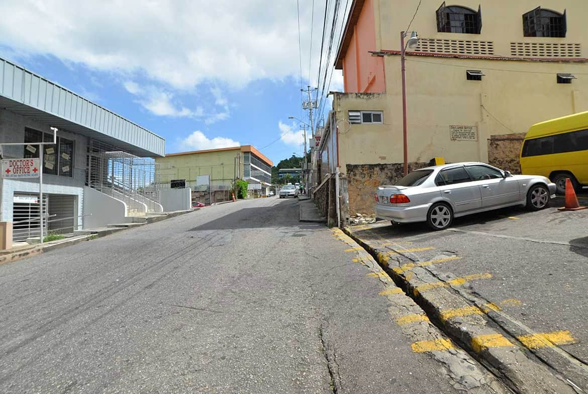 Cornerstone- View of St James Street with parked cars and buildings in Port of Spain.