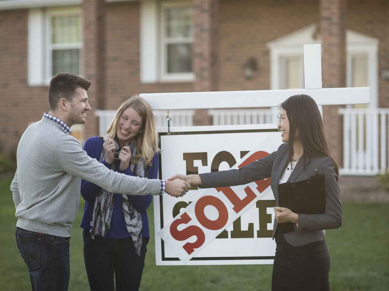 Cornerstone- Happy couple shaking hands with real estate agent in front of sold sign.