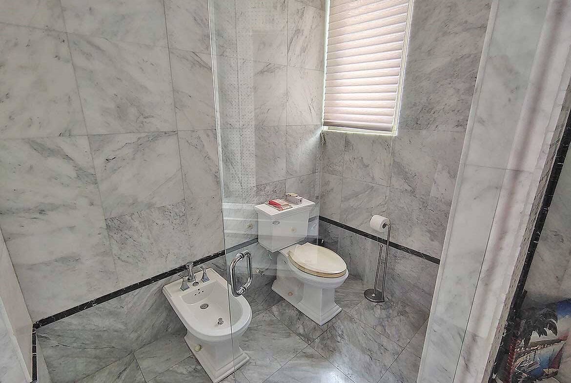 Cornerstone- Contemporary bathroom with marble walls, toilet, bidet, and window with blinds.
