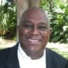 Cornerstone- Smiling man in formal attire outdoors with lush greenery.