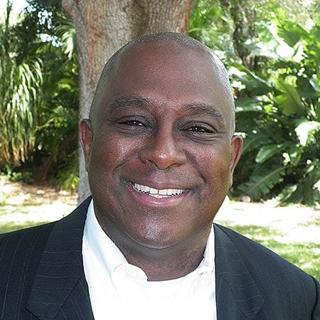 Cornerstone- Smiling man in formal attire outdoors with lush greenery.