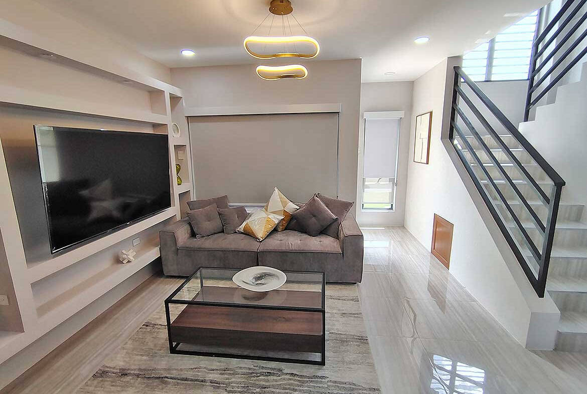 Contemporary living room with gray sofa, wall-mounted TV, and staircase at Ella Vista Gardens.