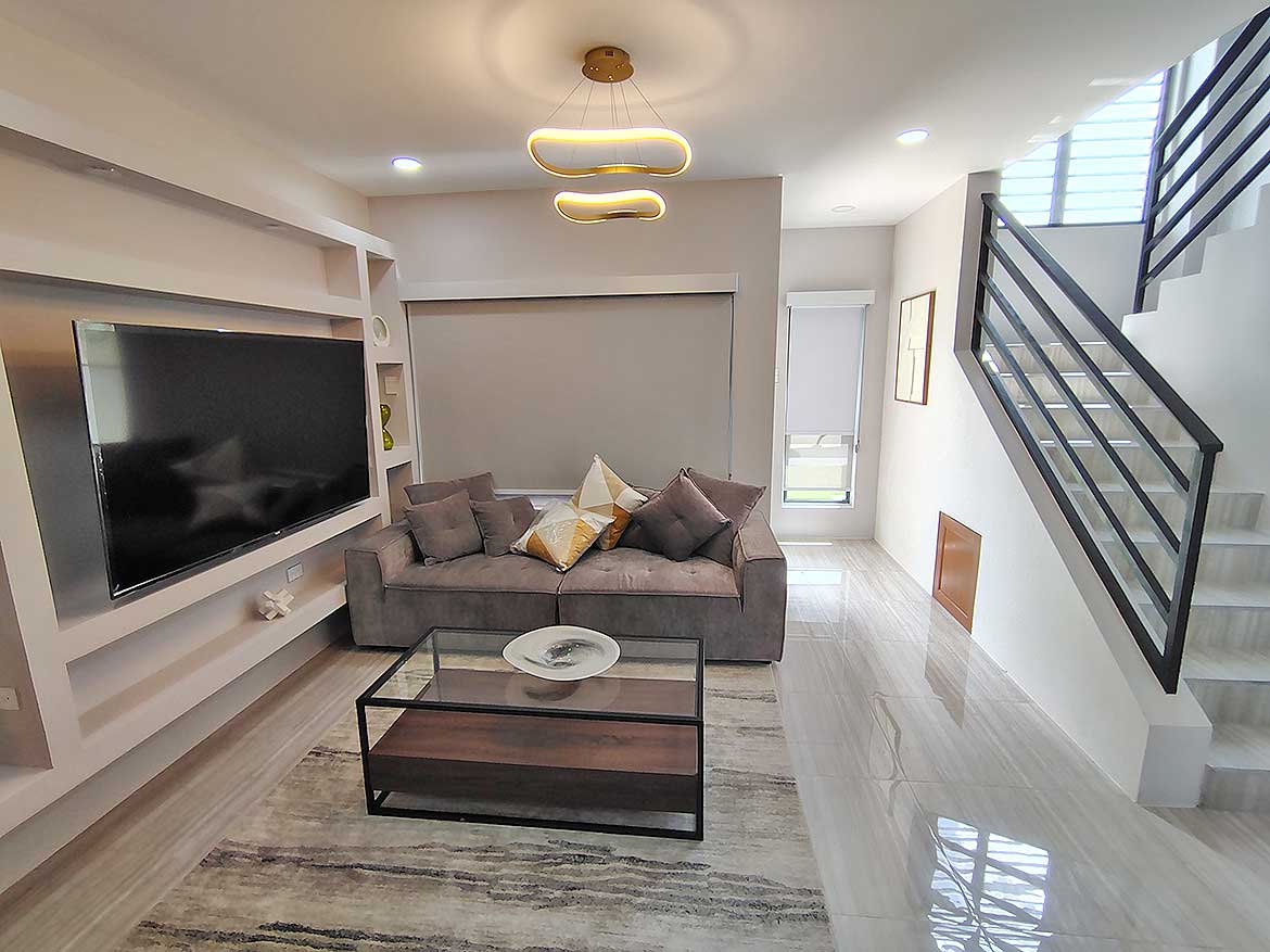 Modern Living Room at Ella Vista Gardens Contemporary living room with gray sofa, wall-mounted TV, and staircase at Ella Vista Gardens.