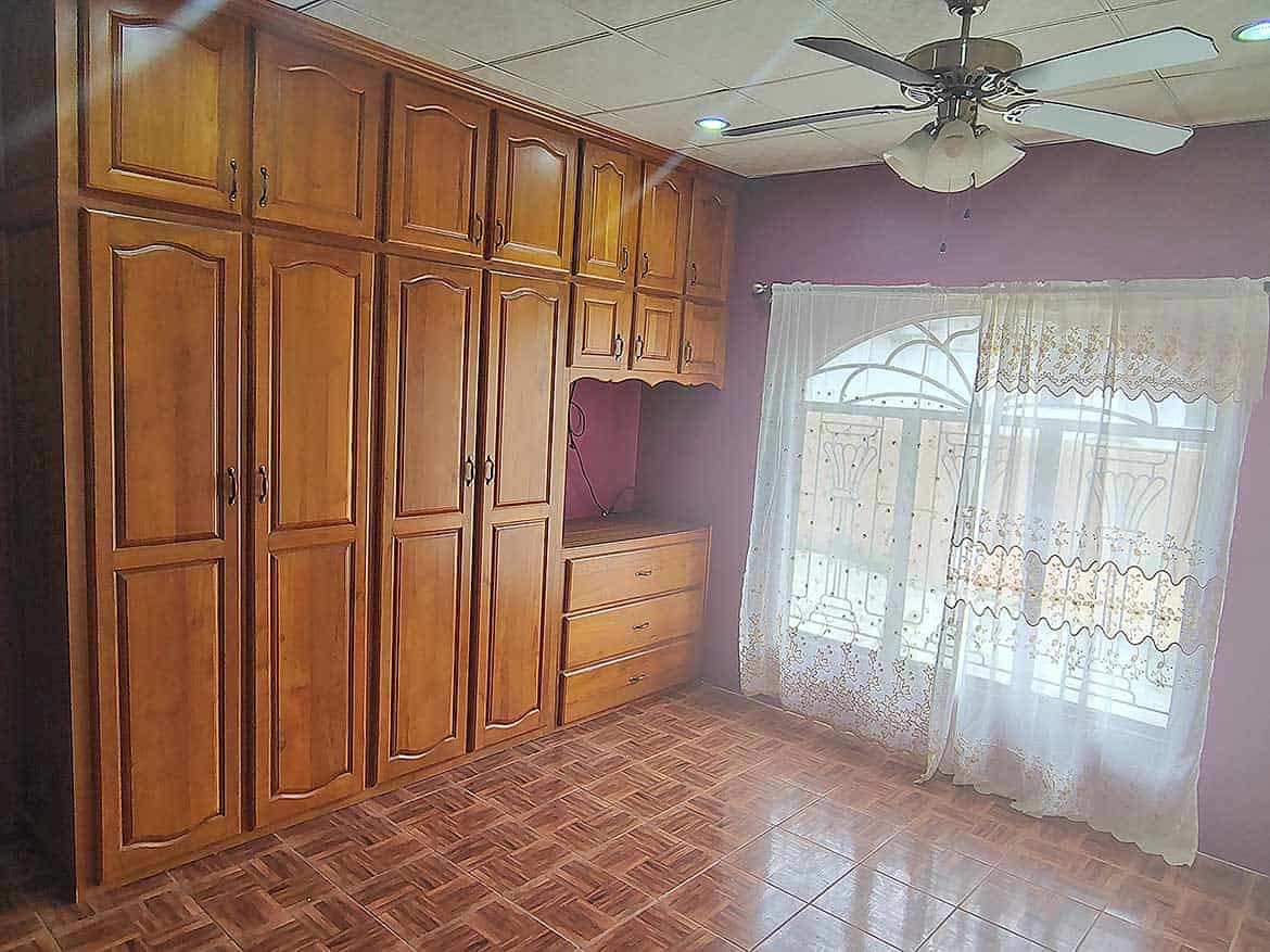 Cornerstone™ Home Interior in Arima Cornerstone- Spacious room with wooden built-in wardrobes and large window with curtains.