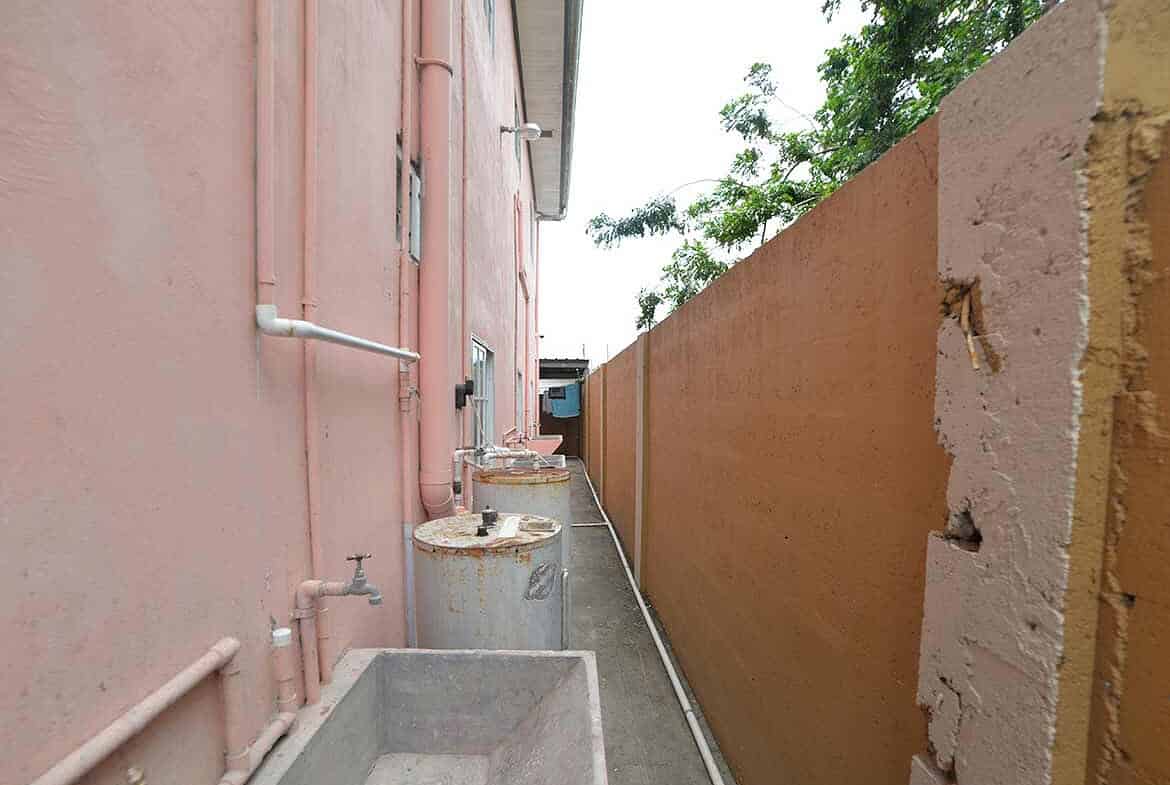 Cornerstone- Narrow alleyway with water tanks and a high wall in Arima.